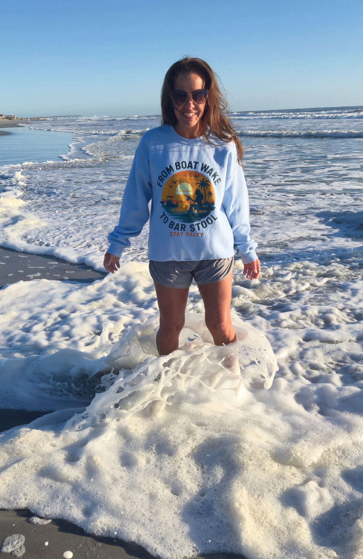 Woman wearing Salty Kiss “From Boat Wake to Bar Stool” coastal sweatshirt while standing in ocean surf on a Florida beach