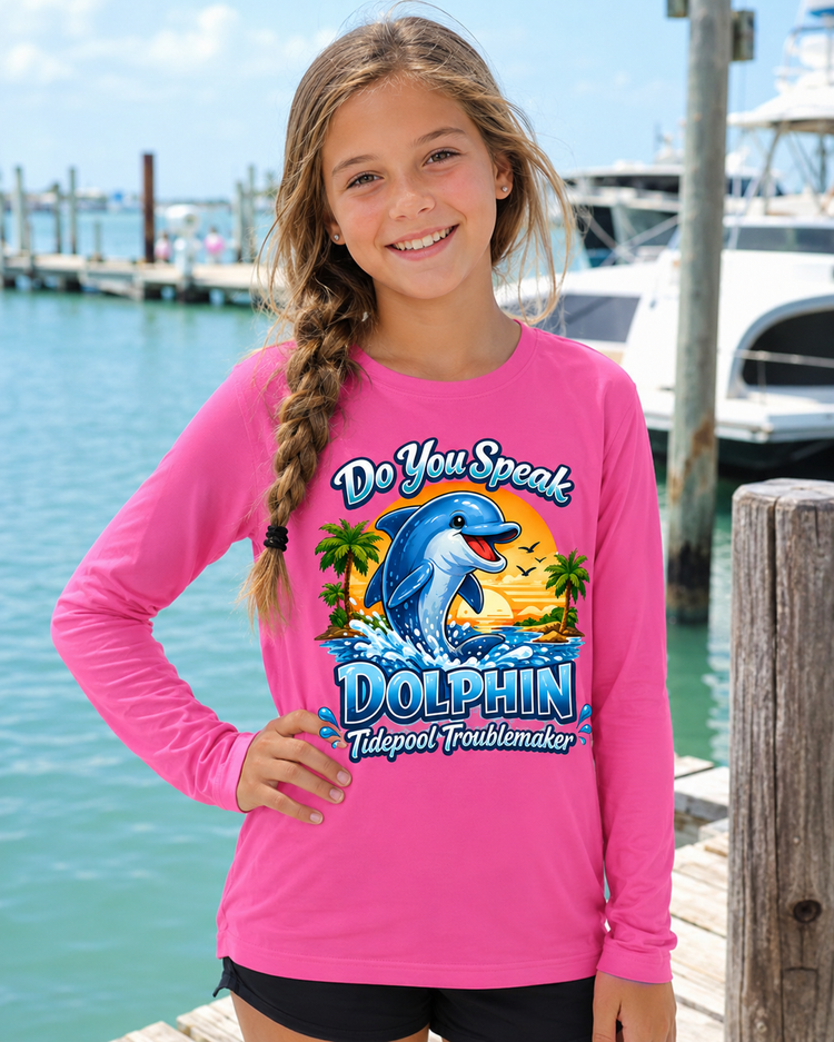 Youth girl wearing a hot pink Tidepool Troublemaker “Do You Speak Dolphin” long sleeve shirt standing by a marina with boats and water in the background.