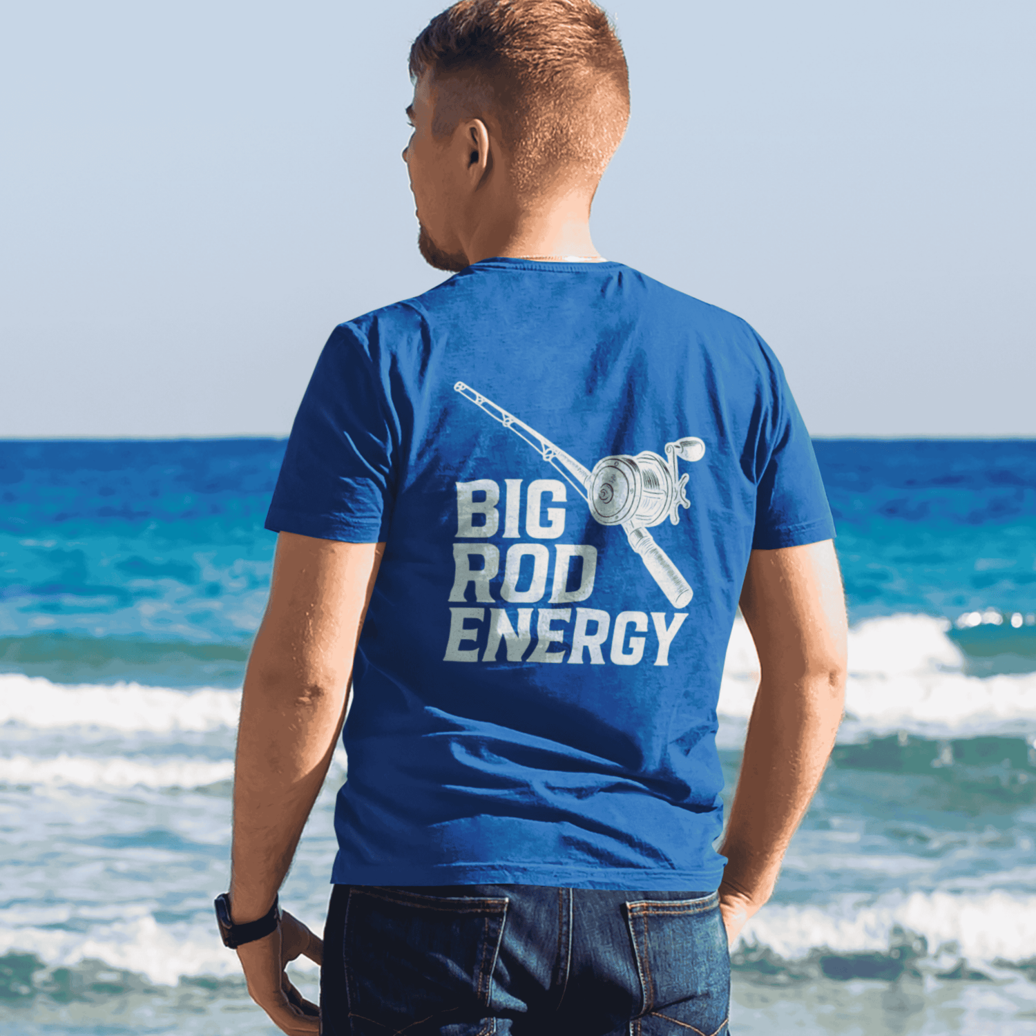 Salty Kiss men’s Big Rod Energy fishing graphic tee worn on a sunny beach with ocean waves.