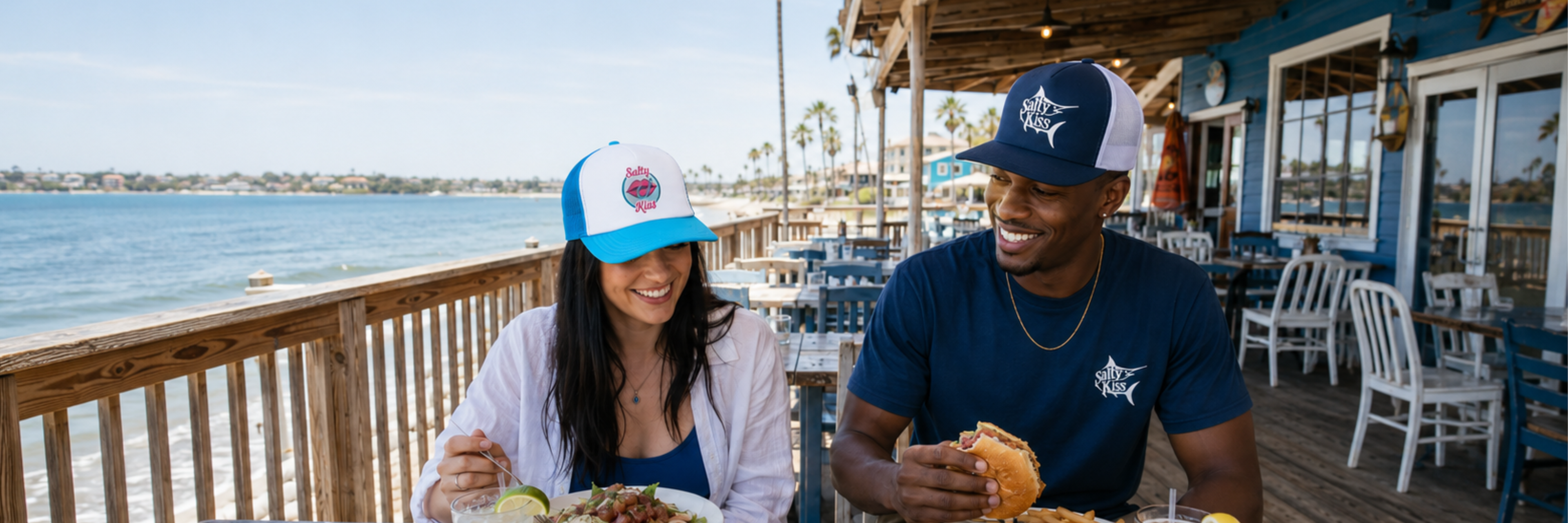 Salty Kiss embroidered coastal hats worn by a man and woman at a waterfront seafood restaurant.
