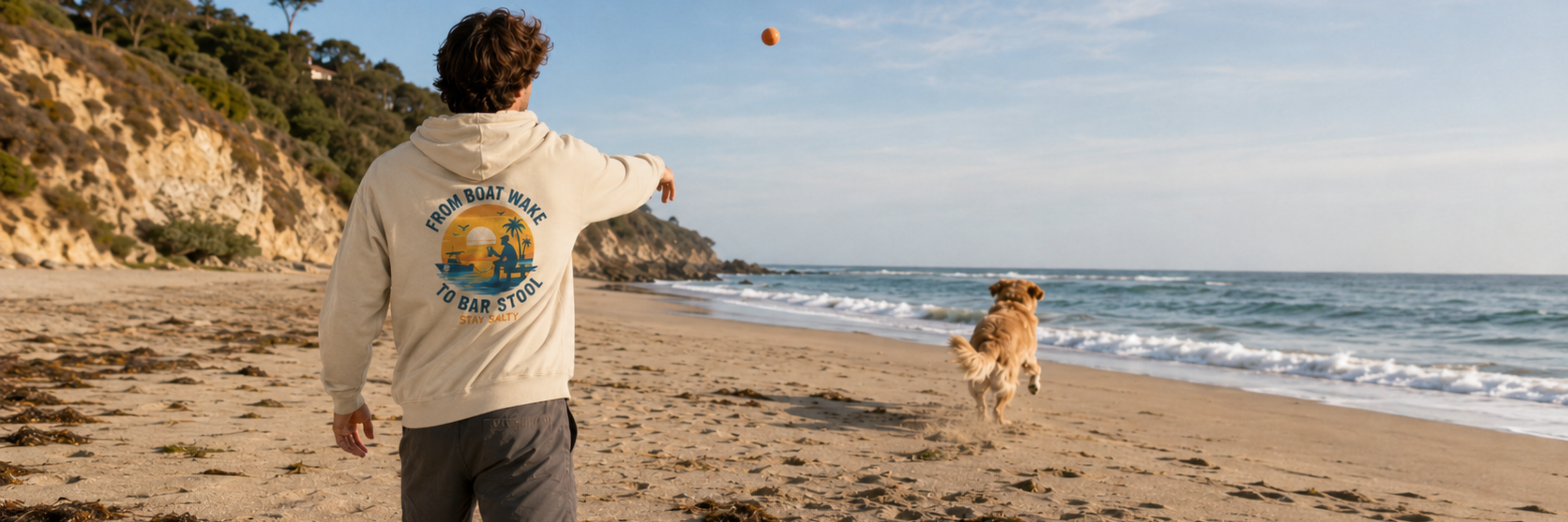 Salty Kiss men’s coastal graphic hoodie worn on a beach while throwing a ball for a dog by the ocean.