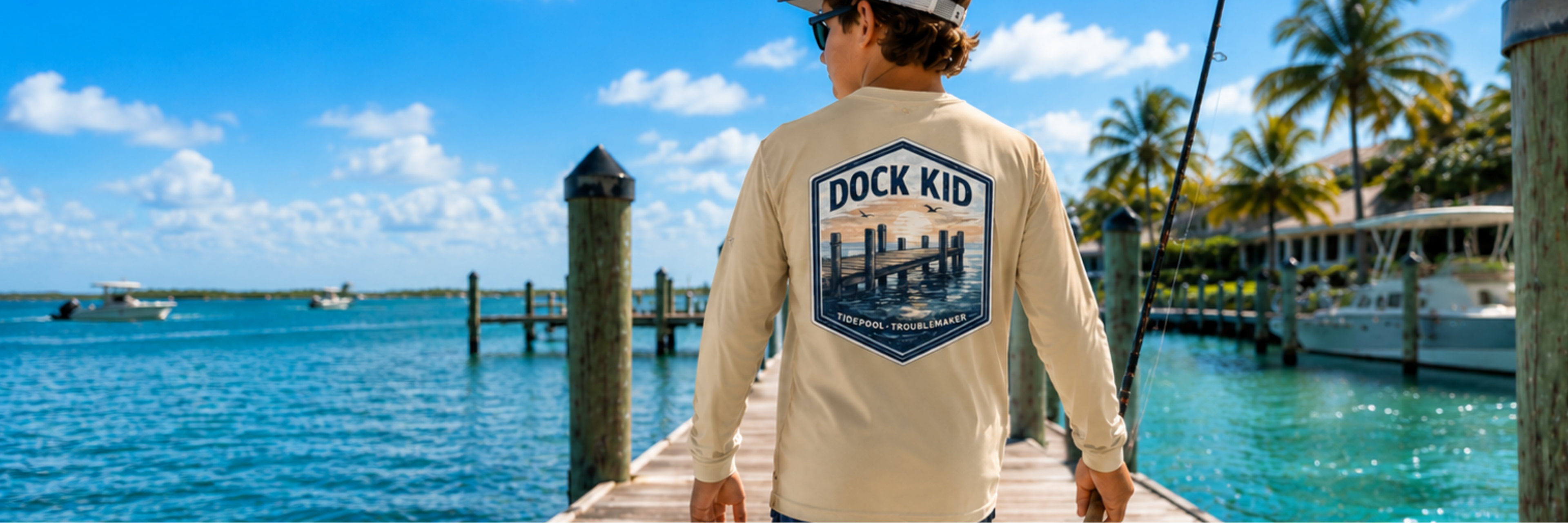 Youth boy wearing a cream Tidepool Troublemaker Dock Kid long sleeve fishing shirt while holding a fishing rod on a sunny coastal dock.